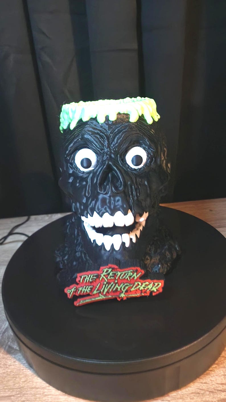 May include: A black, textured zombie head sculpture with bright green hair, wide white eyes, and white teeth. A red and green plaque at the base features the text "THE RETURN OF THE LIVING DEAD" in a stylized font. The figure has a glossy finish.