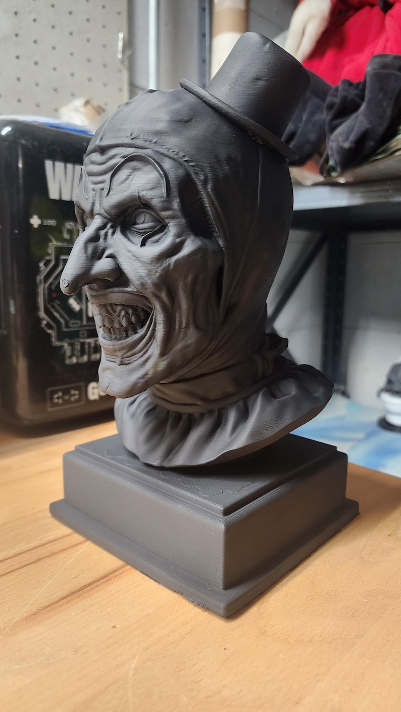 K&ouml;nnte beinhalten: Eine mattschwarze, 3D-gedruckte B&uuml;ste einer Clownfigur mit Zylinder, spitzer Nase und einem breiten, zahnigen Grinsen. Die Figur ist von den Schultern aufw&auml;rts dargestellt, tr&auml;gt einen R&uuml;schenkragen und steht auf einem quadratischen Sockel.