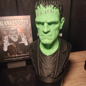 May include: A green-skinned bust of Frankenstein's monster with dark hair, a black jacket, and neck bolts. Beside it stands a Blu-ray case titled Frankenstein Complete Legacy Collection, featuring an image of the monster and the text All 8 Films From The Original Legacy 1931-1948.