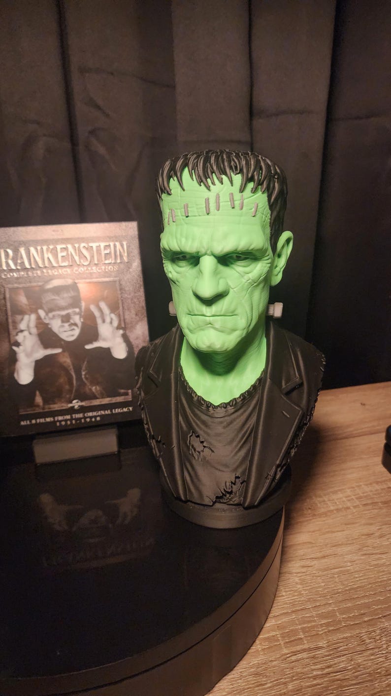 May include: A green-skinned bust of Frankenstein's monster with dark hair, a black jacket, and neck bolts. Beside it stands a Blu-ray case titled Frankenstein Complete Legacy Collection, featuring an image of the monster and the text All 8 Films From The Original Legacy 1931-1948.