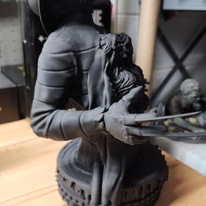 May include: A matte black 3D printed bust of a figure wearing a fedora hat and a striped sweater. The figure has a clawed glove on one hand with long blades extending outward. The bust is mounted on a circular base with a textured, gear-like edge.