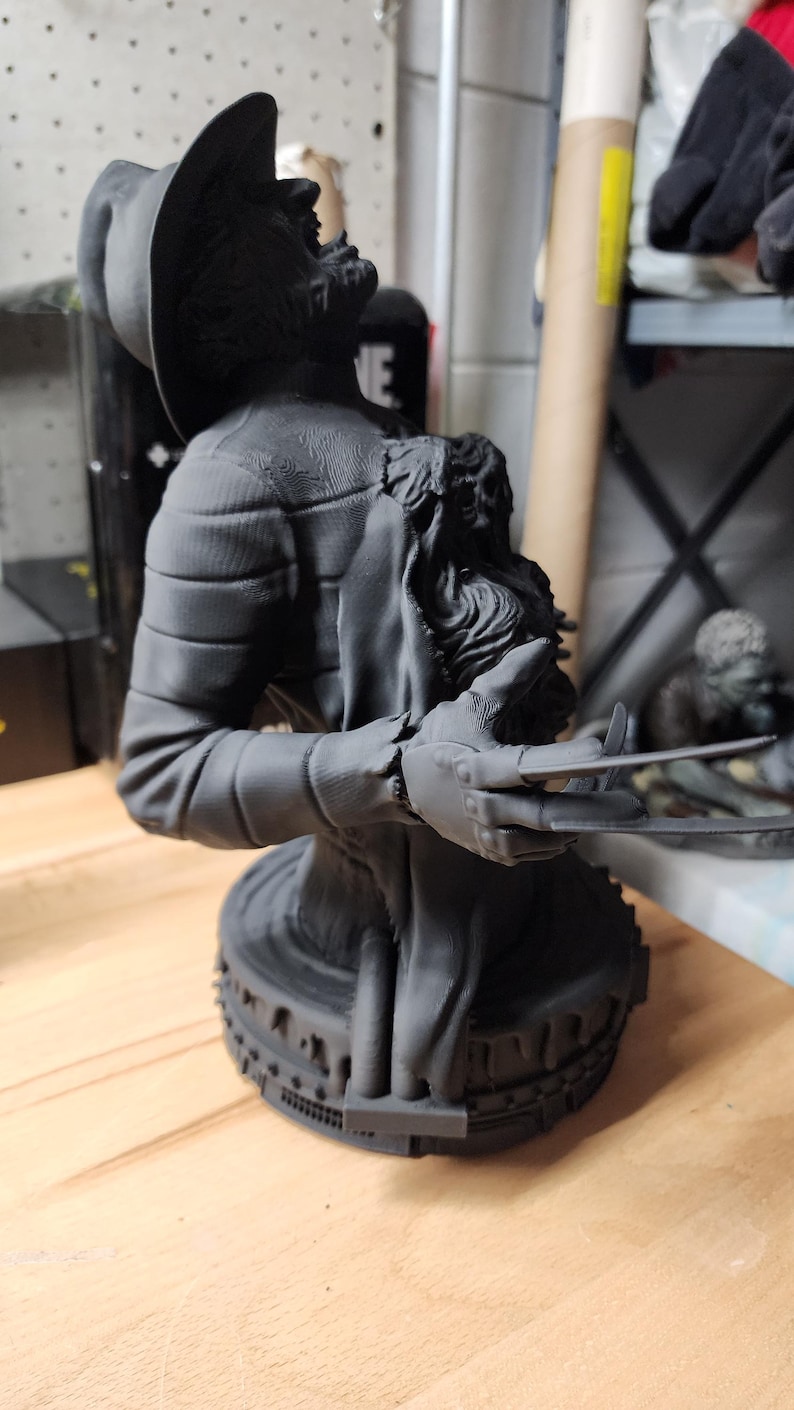May include: A matte black 3D printed bust of a figure wearing a fedora hat and a striped sweater. The figure has a clawed glove on one hand with long blades extending outward. The bust is mounted on a circular base with a textured, gear-like edge.