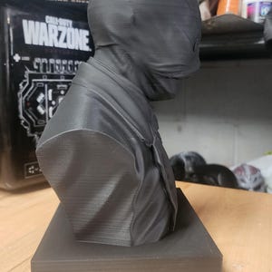 May include: A matte black 3D printed bust of a figure wearing a smooth, featureless mask that covers the entire head and face. The figure is dressed in a collared shirt and tie, mounted on a rectangular base. The surface shows fine horizontal print lines.