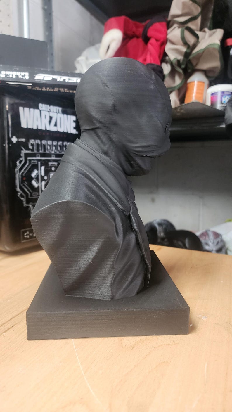 May include: A matte black 3D printed bust of a figure wearing a smooth, featureless mask that covers the entire head and face. The figure is dressed in a collared shirt and tie, mounted on a rectangular base. The surface shows fine horizontal print lines.