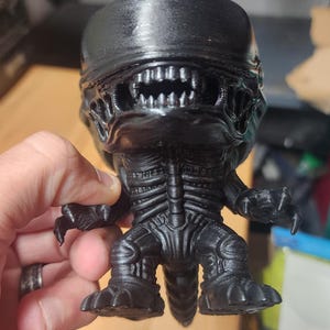 May include: A black 3D-printed figurine of a Xenomorph creature from the Alien franchise. The figure features a smooth, elongated head, a detailed skeletal torso, sharp claws on its hands and feet, and a visible row of teeth within its open mouth.