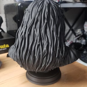 May include: A matte black 3D printed bust sculpture featuring a figure with long, textured, wavy hair that cascades down the shoulders. The sculpture is mounted on a circular pedestal base and is rendered in a solid, monochromatic dark finish.