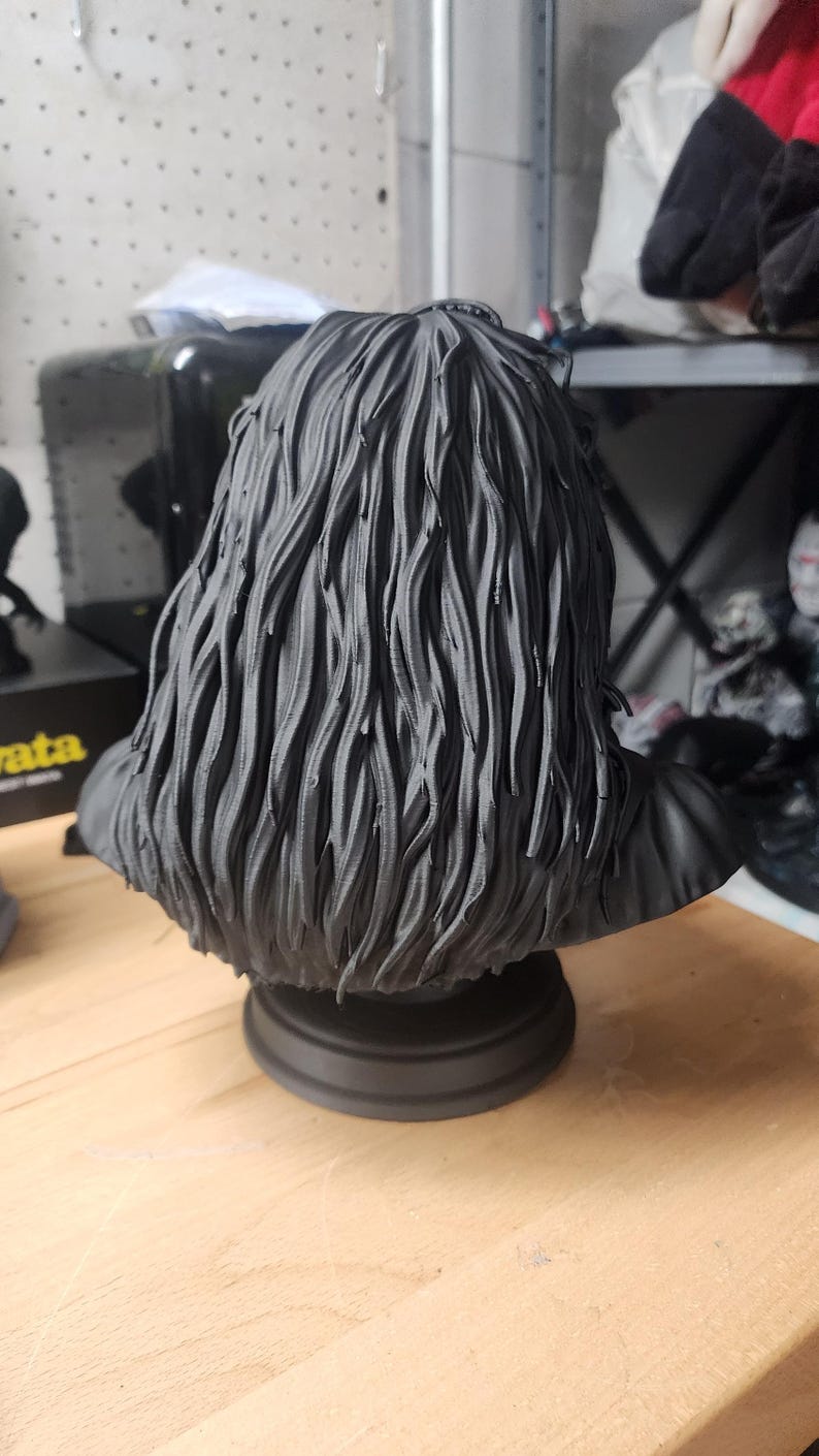 May include: A matte black 3D printed bust sculpture featuring a figure with long, textured, wavy hair that cascades down the shoulders. The sculpture is mounted on a circular pedestal base and is rendered in a solid, monochromatic dark finish.