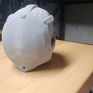 May include: A light gray, 3D-printed helmet-shaped object with a smooth, matte finish. The helmet features a central ridge, a small circular button detail at the top, and a small hole at the base. The surface shows subtle layer lines.