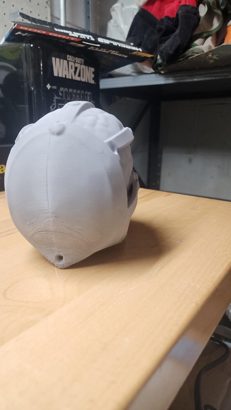 May include: A light gray, 3D-printed helmet-shaped object with a smooth, matte finish. The helmet features a central ridge, a small circular button detail at the top, and a small hole at the base. The surface shows subtle layer lines.
