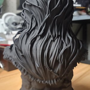 May include: A matte black 3D printed bust sculpture featuring a figure with long, flowing, textured hair that cascades down the back. The base of the sculpture is circular and displays a visible crack running vertically along the front surface.