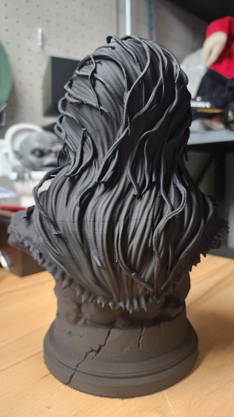 May include: A matte black 3D printed bust sculpture featuring a figure with long, flowing, textured hair that cascades down the back. The base of the sculpture is circular and displays a visible crack running vertically along the front surface.