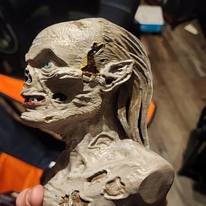 May include: A grey, textured bust of a creature with a gaunt, skeletal face, pointed ears, and long hair. The figure features visible wounds on the temple and chest with dark, glossy accents, and a single visible blue eye set in a deep socket.