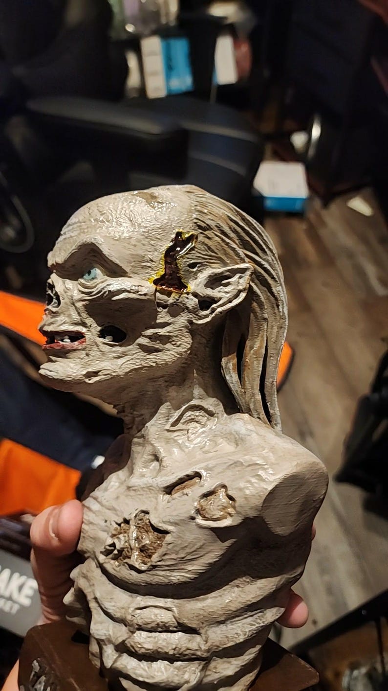 May include: A grey, textured bust of a creature with a gaunt, skeletal face, pointed ears, and long hair. The figure features visible wounds on the temple and chest with dark, glossy accents, and a single visible blue eye set in a deep socket.
