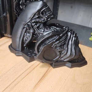 May include: A matte black 3D printed sculpture of a Xenomorph creature from the Alien franchise. The figure features a smooth elongated head, intricate biomechanical ribbing, and a segmented body, all mounted on a flat, contoured base.