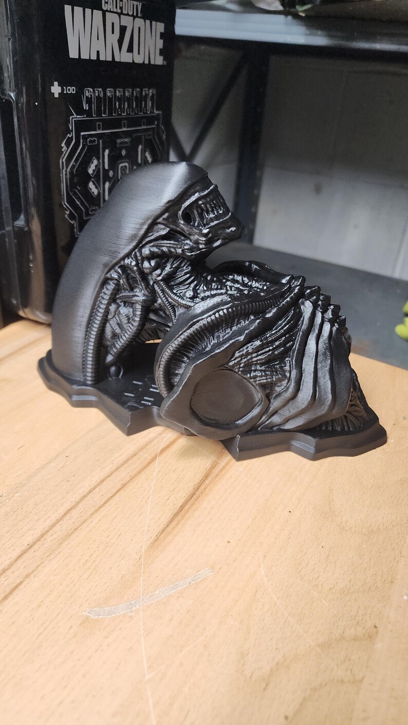 May include: A matte black 3D printed sculpture of a Xenomorph creature from the Alien franchise. The figure features a smooth elongated head, intricate biomechanical ribbing, and a segmented body, all mounted on a flat, contoured base.