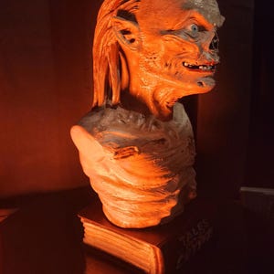 May include: A sculpted bust of a creature with wrinkled skin, pointed ears, and a wide mouth, mounted on a rectangular base shaped like a book with the text Tales from the Crypt visible on the spine, illuminated by a warm orange light.