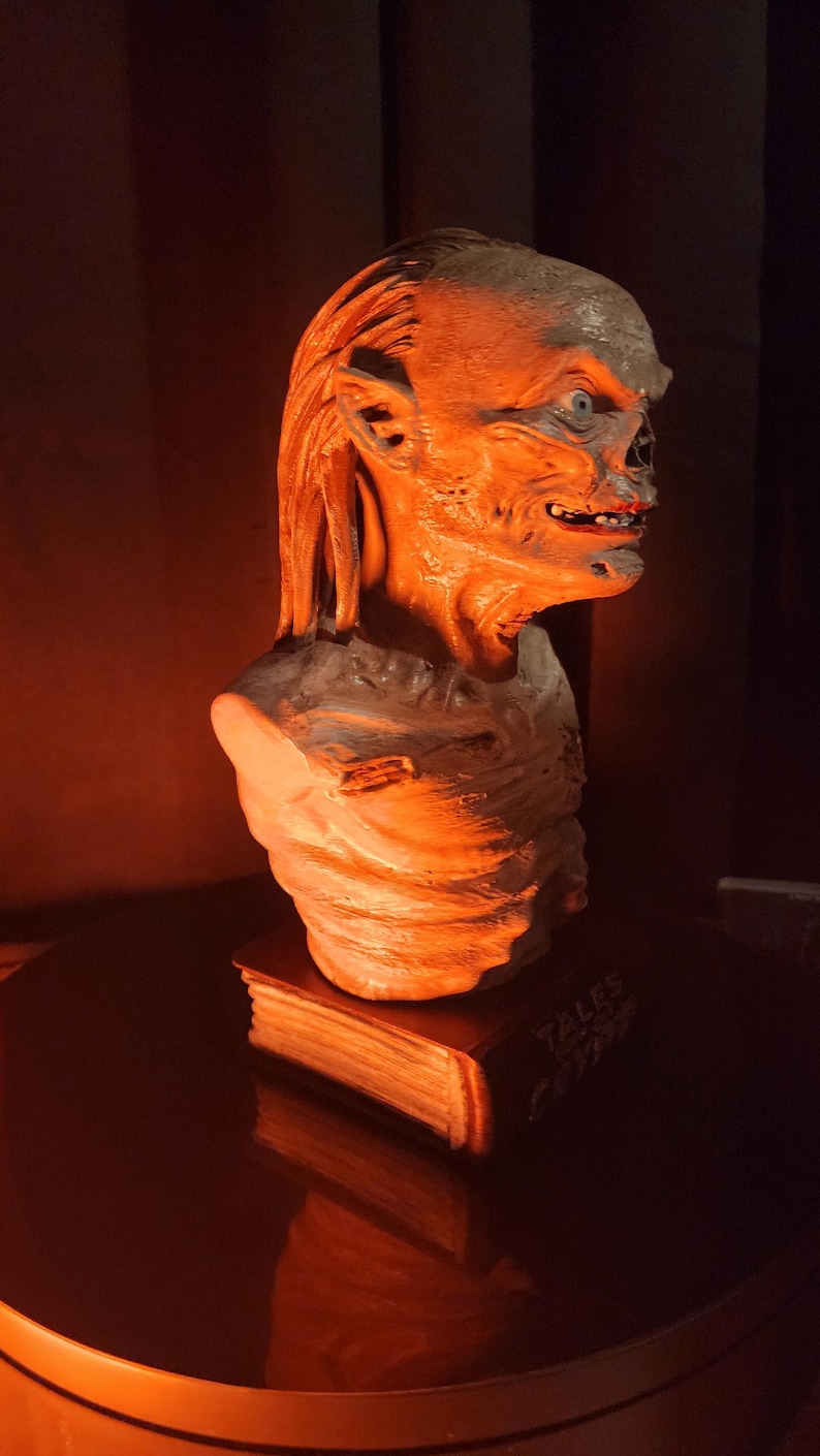 May include: A sculpted bust of a creature with wrinkled skin, pointed ears, and a wide mouth, mounted on a rectangular base shaped like a book with the text Tales from the Crypt visible on the spine, illuminated by a warm orange light.