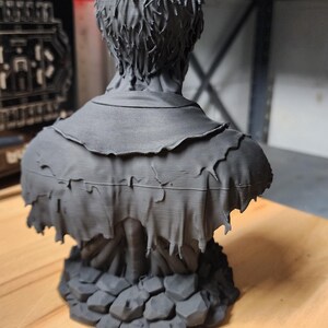 以下が含まれることがあります： 短い質感のある髪をした人物のマットブラックな3Dプリント胸像。端がほつれたボロボロの重ね着風の衣服を身に着けている。彫刻は、不規則に積み重なった石のような形状の台座の上に設置されており、細かいプリントの積層跡が見える。
