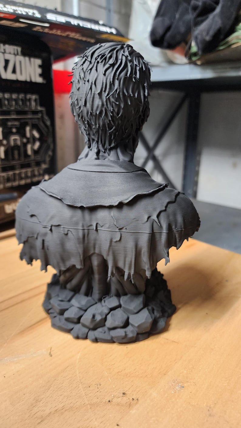 以下が含まれることがあります： 短い質感のある髪をした人物のマットブラックな3Dプリント胸像。端がほつれたボロボロの重ね着風の衣服を身に着けている。彫刻は、不規則に積み重なった石のような形状の台座の上に設置されており、細かいプリントの積層跡が見える。
