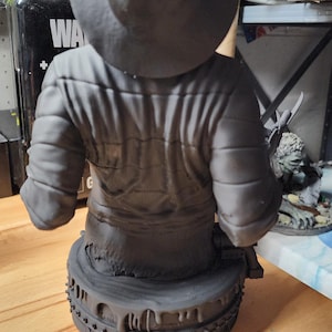 May include: A matte gray resin bust of a figure wearing a wide-brimmed hat and a quilted jacket. The figure is positioned on a circular base featuring a textured, dripping design with small rectangular details along the lower edge of the base.