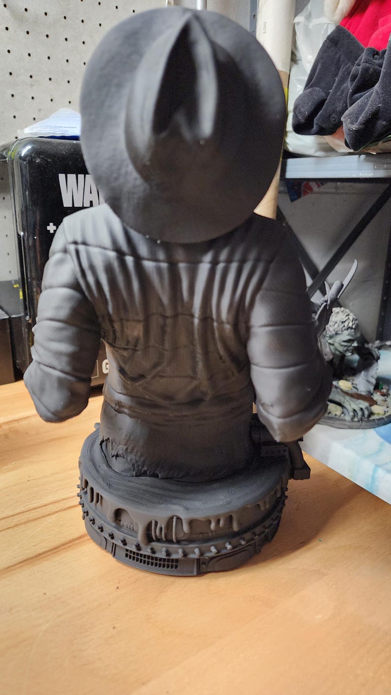 May include: A matte gray resin bust of a figure wearing a wide-brimmed hat and a quilted jacket. The figure is positioned on a circular base featuring a textured, dripping design with small rectangular details along the lower edge of the base.