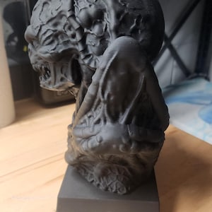 Puede incluir: Un busto negro mate impreso en 3D de una criatura con una superficie org&aacute;nica texturizada. La figura presenta una cabeza redondeada con patrones intrincados similares a venas, una boca gru&ntilde;endo con dientes visibles y una base rectangular lisa.