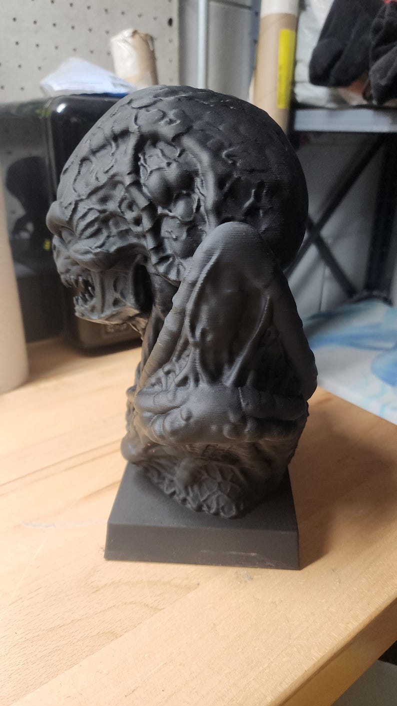 Puede incluir: Un busto negro mate impreso en 3D de una criatura con una superficie org&aacute;nica texturizada. La figura presenta una cabeza redondeada con patrones intrincados similares a venas, una boca gru&ntilde;endo con dientes visibles y una base rectangular lisa.