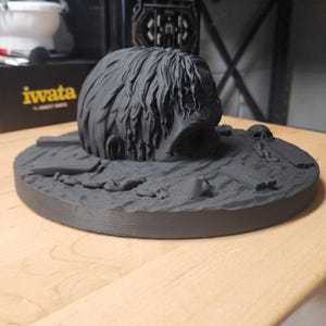 May include: A matte black 3D printed sculpture featuring a human head emerging from a textured ground surface. The base includes small sculpted details such as a skull, debris, and planks. The head has detailed hair texture and a wide open mouth.