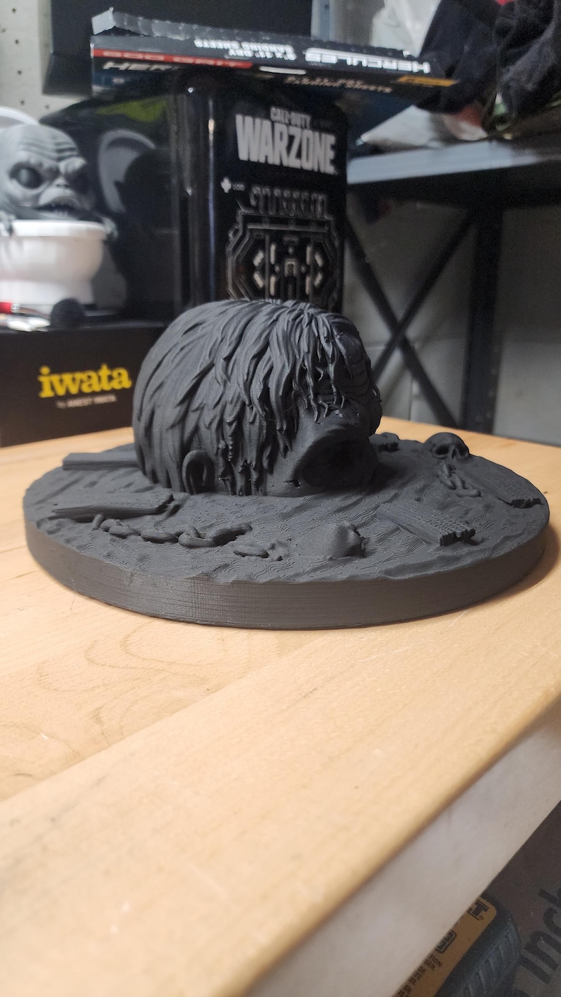 May include: A matte black 3D printed sculpture featuring a human head emerging from a textured ground surface. The base includes small sculpted details such as a skull, debris, and planks. The head has detailed hair texture and a wide open mouth.