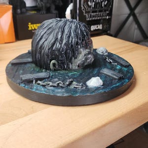 May include: A 3D printed sculpture featuring a dark, textured head emerging from a circular base painted with blue and green water effects. The base includes small sculpted details like wooden planks, metal chains, and white skull-like accents.