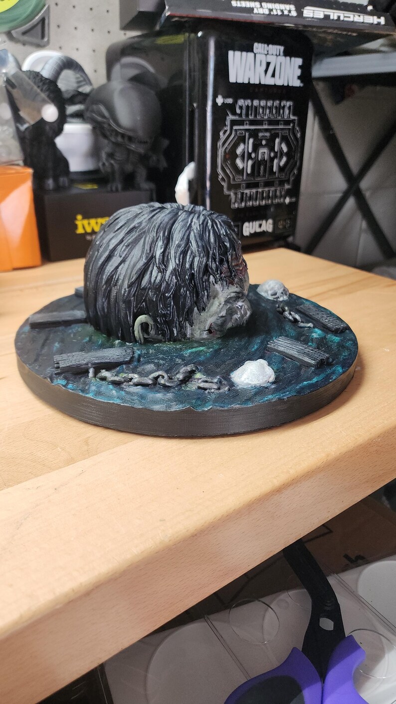 May include: A 3D printed sculpture featuring a dark, textured head emerging from a circular base painted with blue and green water effects. The base includes small sculpted details like wooden planks, metal chains, and white skull-like accents.