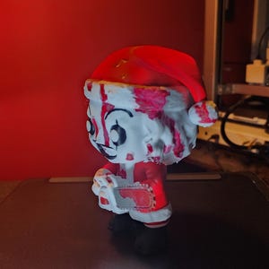 May include: A stylized vinyl figure of a character wearing a red and white Santa hat and suit, covered in red blood-like splatters. The figure has a pale face with dark eye makeup and holds a small, blood-stained chainsaw in its hands.