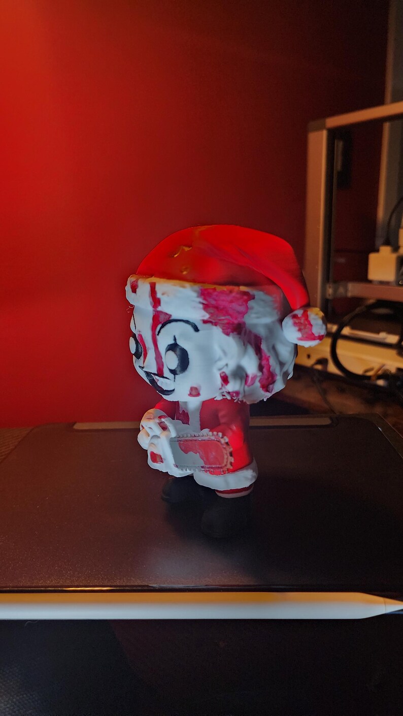 May include: A stylized vinyl figure of a character wearing a red and white Santa hat and suit, covered in red blood-like splatters. The figure has a pale face with dark eye makeup and holds a small, blood-stained chainsaw in its hands.