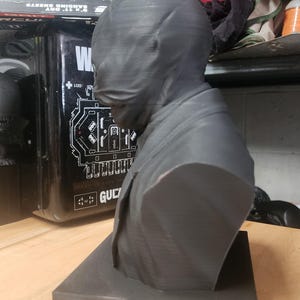 May include: A dark grey 3D-printed bust of a figure wearing a smooth, featureless mask that covers the entire head and face. The figure is dressed in a collared jacket and is mounted on a square, flat base, showcasing visible horizontal print lines.
