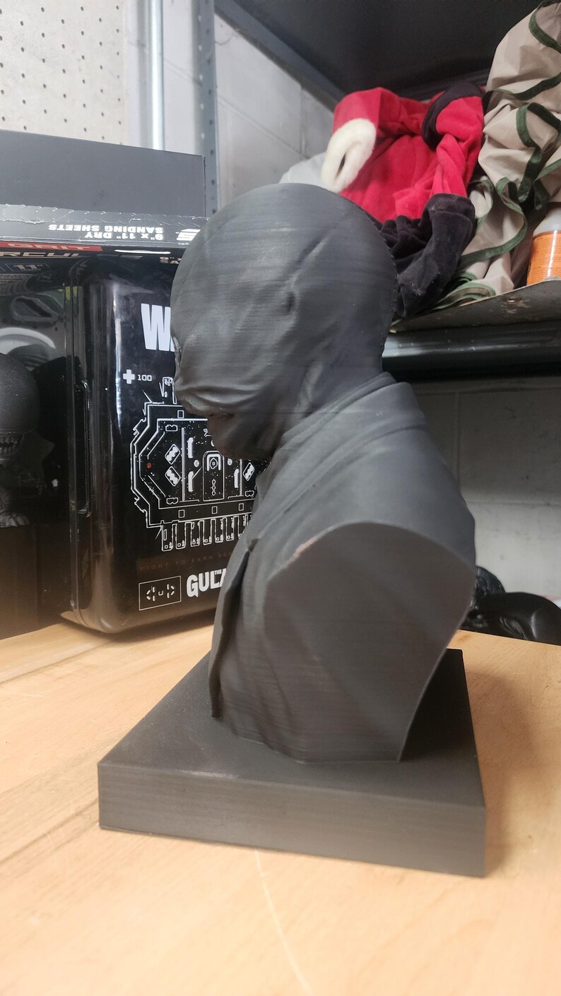 May include: A dark grey 3D-printed bust of a figure wearing a smooth, featureless mask that covers the entire head and face. The figure is dressed in a collared jacket and is mounted on a square, flat base, showcasing visible horizontal print lines.