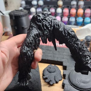 May include: A black 3D-printed miniature terrain piece featuring a jagged, arching structure with a textured, organic surface. The object is matte black and displays visible layer lines from the printing process, with a separate circular base.