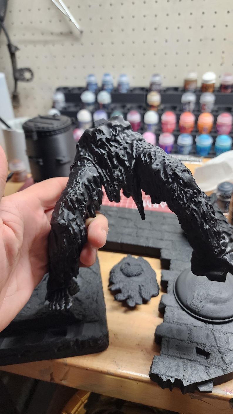 May include: A black 3D-printed miniature terrain piece featuring a jagged, arching structure with a textured, organic surface. The object is matte black and displays visible layer lines from the printing process, with a separate circular base.