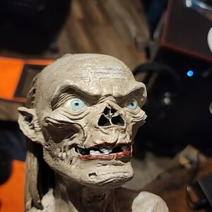 May include: A sculpted bust of a creature with pale, textured skin, prominent facial features, and bright blue eyes. The figure has a wide, toothy grin with dark red lips, pointed ears, and a gaunt, skeletal appearance with visible muscle definition.