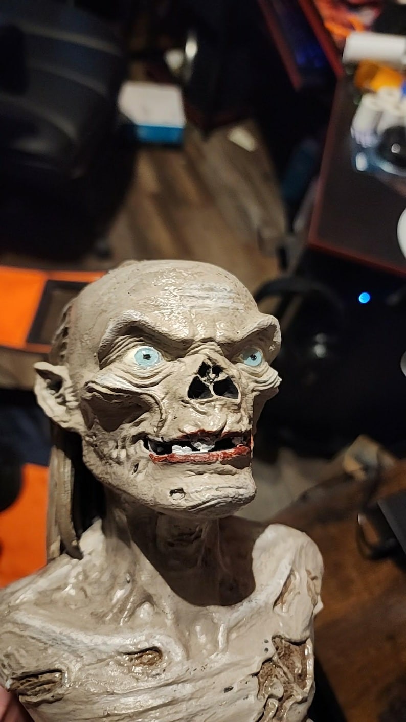 May include: A sculpted bust of a creature with pale, textured skin, prominent facial features, and bright blue eyes. The figure has a wide, toothy grin with dark red lips, pointed ears, and a gaunt, skeletal appearance with visible muscle definition.