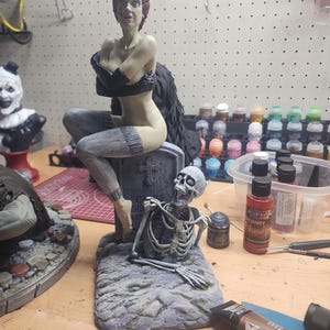 Puede incluir: Una figura de resina pintada que muestra a un personaje con cabello rojo, guantes negros y medias grises sentado sobre una l&aacute;pida gris. Un esqueleto blanco emerge del suelo frente a la l&aacute;pida. La base tiene una textura que imita la piedra.