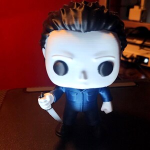 May include: A stylized vinyl figure of a character with a white mask, black hair, and a dark blue jumpsuit. The figure has large black circular eyes and is holding a small white knife in its right hand. The design features a large head and a small body.