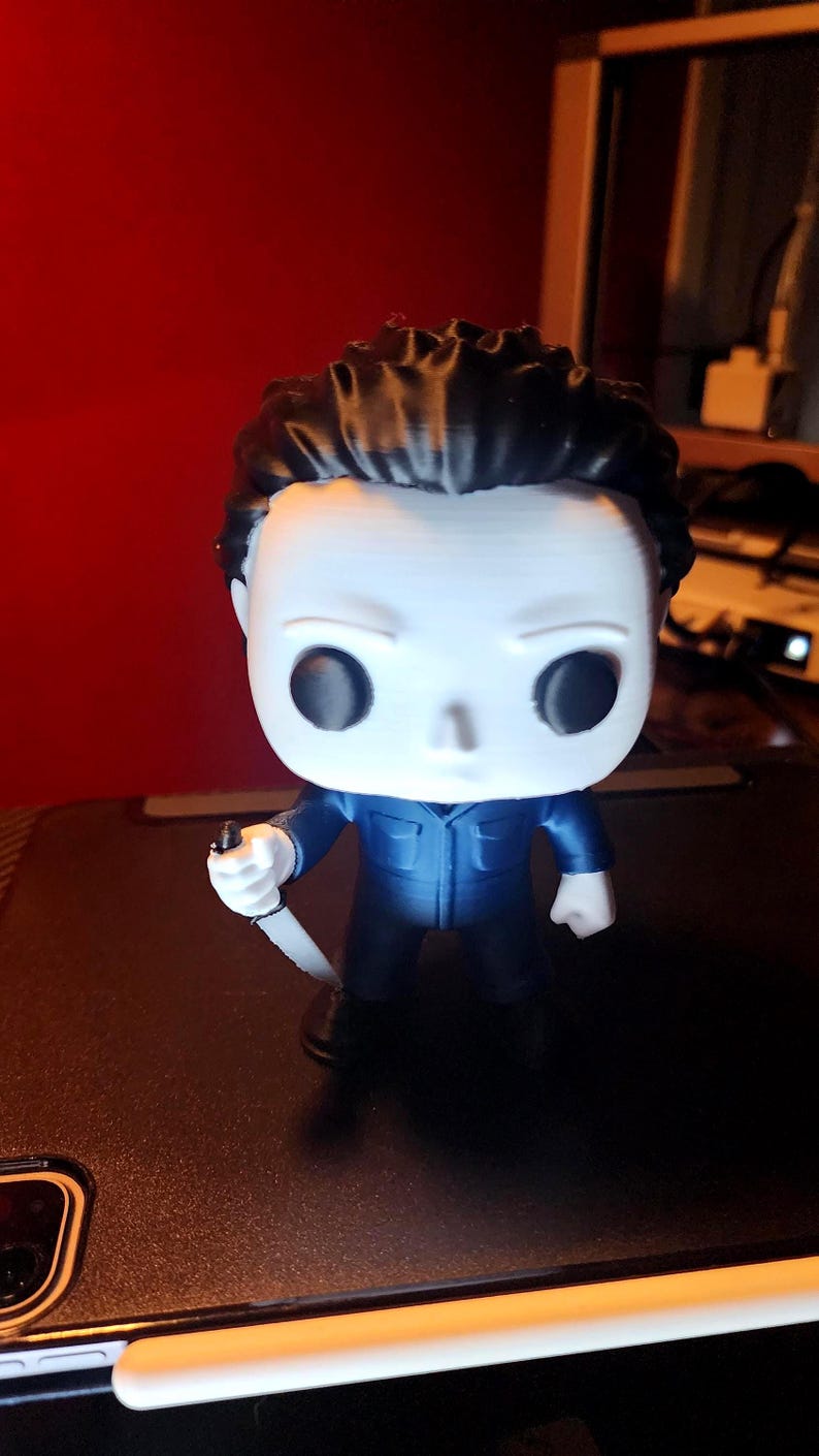 May include: A stylized vinyl figure of a character with a white mask, black hair, and a dark blue jumpsuit. The figure has large black circular eyes and is holding a small white knife in its right hand. The design features a large head and a small body.