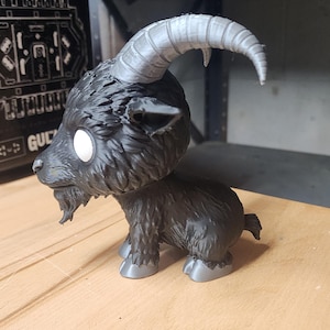 May include: A black goat figurine with a large head, white circular eyes, and a small beard. The goat features prominent, curved, light gray horns and matching light gray hooves. The textured fur is rendered in a dark black finish throughout.