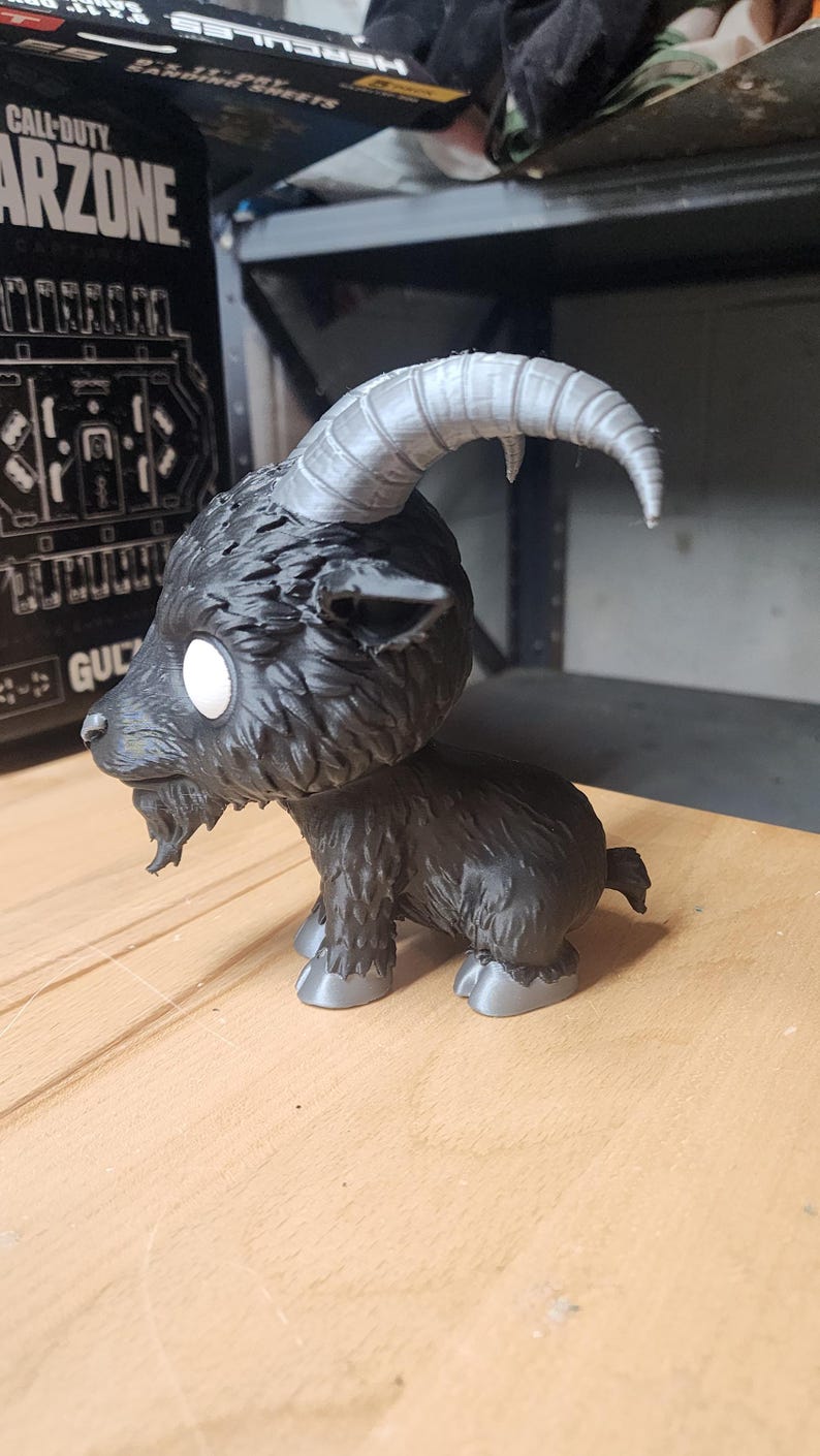 May include: A black goat figurine with a large head, white circular eyes, and a small beard. The goat features prominent, curved, light gray horns and matching light gray hooves. The textured fur is rendered in a dark black finish throughout.