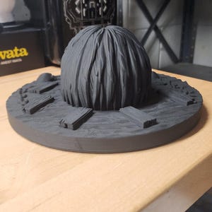 May include: A matte black 3D printed circular display base featuring a central textured dome resembling hair or foliage, surrounded by small rectangular plank-like details arranged radially on a wood-grain patterned surface.