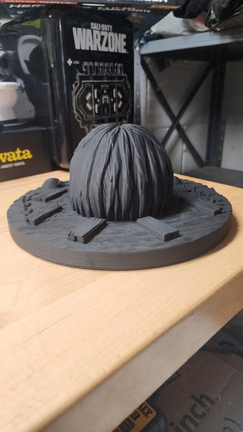 May include: A matte black 3D printed circular display base featuring a central textured dome resembling hair or foliage, surrounded by small rectangular plank-like details arranged radially on a wood-grain patterned surface.