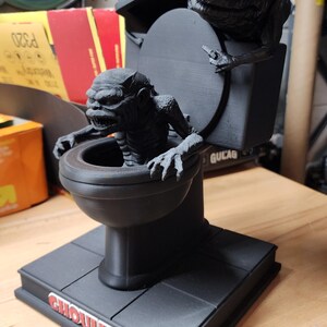 May include: A matte black 3D printed figurine of a toilet with two textured, monstrous creatures emerging from it. One creature sits inside the bowl, while the other perches on the lid. The base features the red and white text GHOULIES.