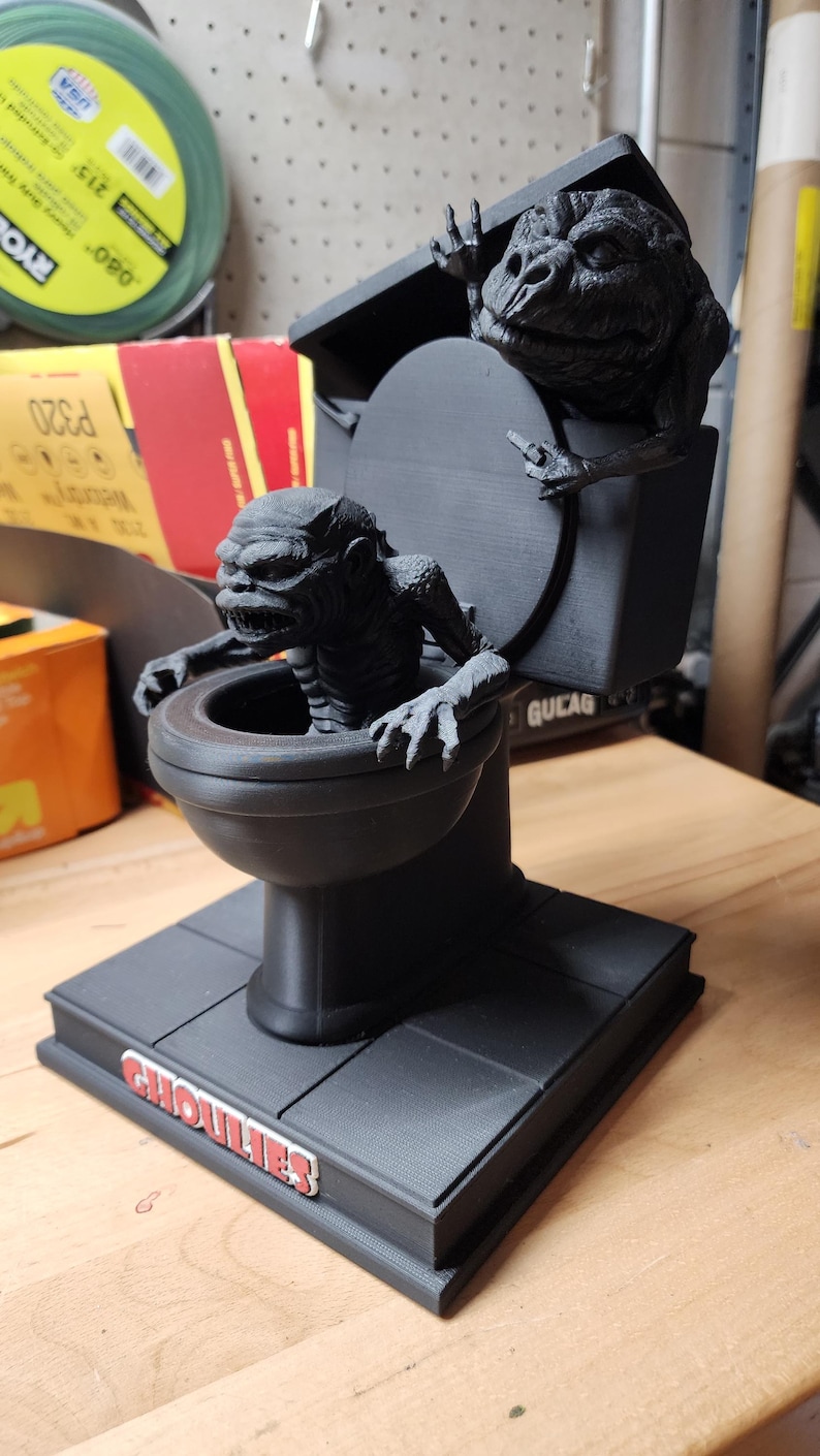 May include: A matte black 3D printed figurine of a toilet with two textured, monstrous creatures emerging from it. One creature sits inside the bowl, while the other perches on the lid. The base features the red and white text GHOULIES.