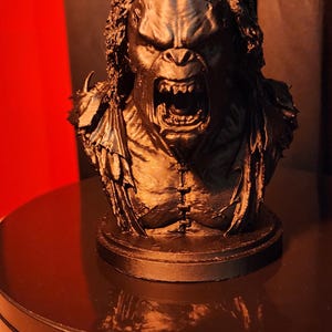 May include: A black monochromatic bust of a snarling creature with textured hair, sharp teeth, and a visible chest seam. The figure features a wide, open mouth and detailed facial expressions, mounted on a circular base, capturing a dark, aggressive aesthetic.