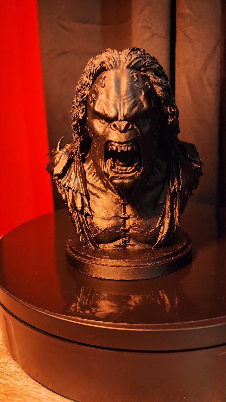 May include: A black monochromatic bust of a snarling creature with textured hair, sharp teeth, and a visible chest seam. The figure features a wide, open mouth and detailed facial expressions, mounted on a circular base, capturing a dark, aggressive aesthetic.